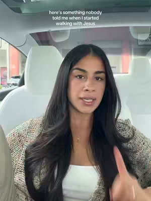 A woman with long black hair is in the front seat of a car, looking directly at the camera and talking. She is wearing a white top under a beige cardigan, and the background shows the car's interior and a view of a building.