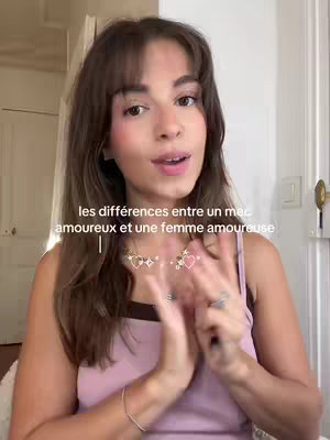 A young woman with long brown hair is positioned in front of a soft-focused background. She wears a light pink top and has a delicate necklace with hearts and stars on. Text overlay across the image says the different between a man in love and a woman in love.
