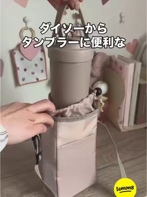 The cover image shows a person holding a light pink tote bag with a pocket on the front. The bag is positioned against a wall featuring a heart decoration and other items. The environment is indoors, suggesting a home setting. The overall color scheme is pastel, and the lighting appears to be soft. The bag appears to be holding a tumbler.