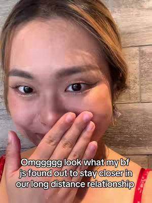 A close-up shot of a person's face, appearing to be on the verge of tears or emotional, with a hand covering the mouth. The person has dark eyes, fair skin, and light-brown hair. In the background, there is slightly textured wall. Text overlayed across the bottom of the image indicates the subject's boyfriend found a way to stay closer in their long distance relationship.