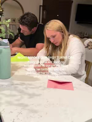 The image shows a woman and a man sitting at a table writing in birthday cards. A mirror is visible in the background, along with decorative plants in the corner of the room. A pink envelope sits on the table.