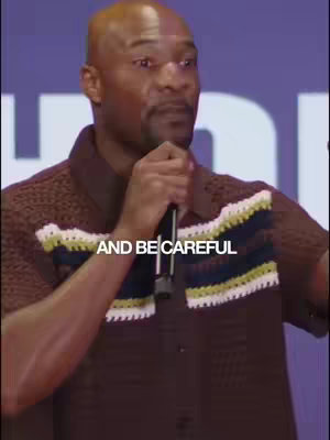 A man in a brown knit shirt holding a microphone, looking toward the left. White text overlayed across the center of the image.