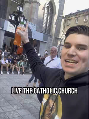 A selfie-style video cover image shows a young man with a bright smile holding a bottle of orange liquid in front of a church building. The man is wearing a black hoodie, and a religious image is visible on his chest. The background includes a crowd of people and various architectural details.