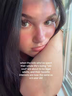 A selfie of a woman looking pensively at the camera, with one hand supporting her head. The background is soft and blurry, with some out-of-focus window visible on the right. Text overlay is visible.