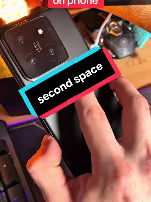 A close-up shot showcasing a black smartphone, positioned at a slight angle, indicating a tech-focused video. The phone’s camera module with the Leica logo is in focus, suggesting the video might review camera features. A hand is holding the phone. A text overlay in a rectangular box with a red and blue outline reading "second space" is a prominent feature. The background includes a wooden surface and a keyboard, offering hints about the setting.