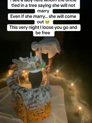 The image shows a medium shot of a person carrying a tray on their head. The person is seen from the back, wearing a patterned shirt and a yellow skirt. Another person, wearing a white outfit, is beside them. The background is dark, with candles creating a warm glow.