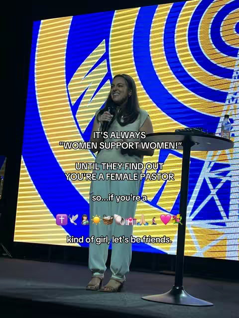 A woman stands on a stage with a microphone, addressing an audience. She's dressed in a casual outfit against a backdrop of yellow and blue abstract shapes. Text is overlaid on the image conveying a message about female support and friendship.