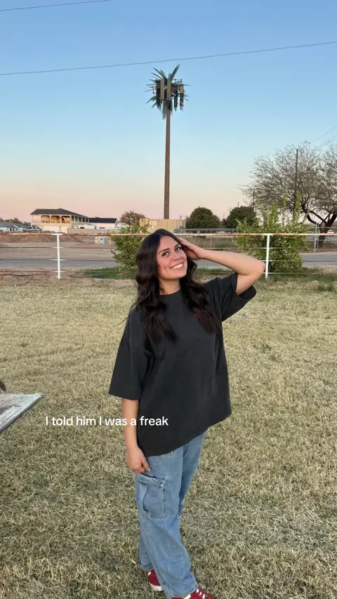A woman with long dark hair smiles while standing on a grassy lawn. She wears a large dark t-shirt and jeans. In the background is a palm tree-shaped cell phone tower.