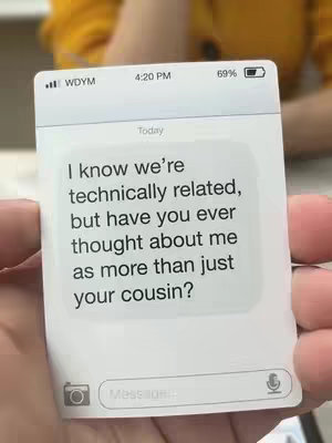 A person's hand holds a white card with a mockup iPhone or text message interface displayed on it. The screen shows a text message conversation with the text on the screen reading, "I know we're technically related, but have you ever thought about me as more than just your cousin?" The top of the phone display reads, "WDYM" with a battery percentage and time stamp. The person holding the phone appears to be wearing a yellow shirt.