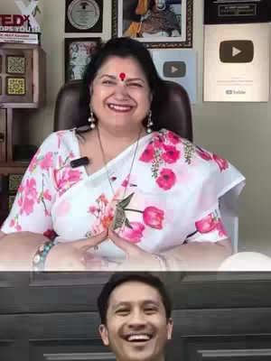 The video cover image features a split screen. The top half shows a woman in a bright floral patterned saree, smiling while seated, with her hands together in a prayer or greeting gesture. The background includes framed pictures and awards, suggesting a professional setting. The bottom half captures a smiling man.