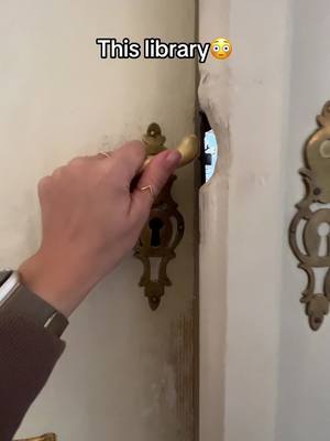 A hand is shown grasping a gold-colored door handle, pulling open a door. The door appears to be aged, with an elaborate escutcheon plate on the right. There is a glimpse of an interior space through the opening, suggesting a library.
