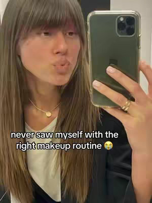 A woman with long, light brown hair and bangs is taking a selfie, holding a phone in the right hand, and pouting at the camera, next to a background wall. She is wearing a white shirt and gold necklace. The image has a warm and neutral color palette. Overlayed text is at the bottom.