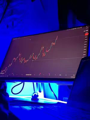 A computer monitor displaying a stock chart with red and green lines against a dark background, lit from below with a cool blue light. A laptop and other objects are visible along the bottom of the frame.