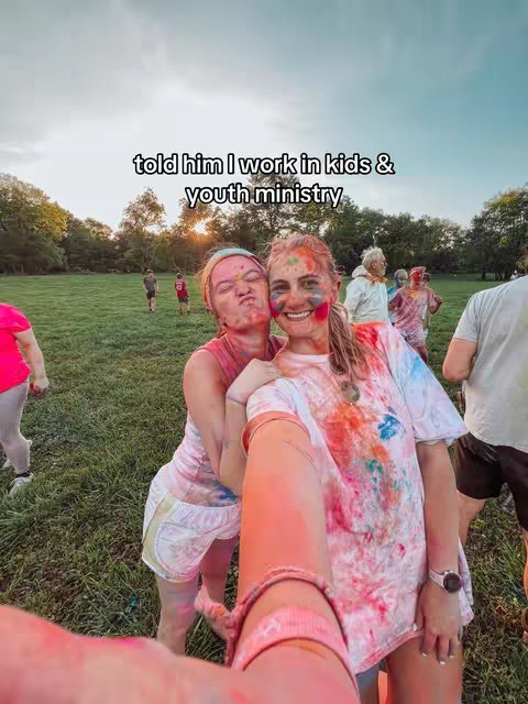Two young women are taking a selfie outdoors with colorful paint on their faces and clothes in a grassy park setting. The image has a slightly overexposed, warm tone, typical of a sunny day. In the background, other people are also covered in paint, suggesting a group activity, likely a color run or festival. The image is composed as a selfie, with the arms of the person taking the photo visible in the foreground.