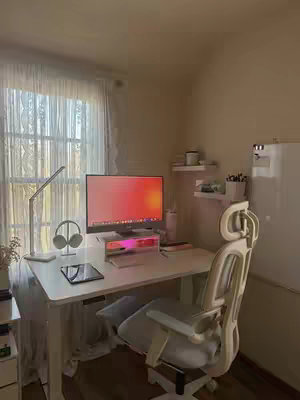 A well-lit work from home setup featuring a white desk, a large monitor displaying a fiery gradient, and a comfortable white office chair. Natural light streams in from a window on the left, illuminating the space. There are other small accessories like headphones and a tablet or book visible as well.