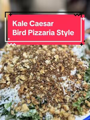 A close-up shot showcasing a salad, likely a Caesar salad with kale and croutons. The salad is topped with crumbled ingredients and a dressing. Overlayed on the image is a red rectangular shape containing text, likely the title of the dish.