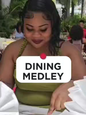 A medium shot shows a woman sitting at a table, appearing to be at a restaurant. She is wearing a green top and has dark hair with a styled fringe. A white rectangular text overlay appears centered, with the words "DINING MEDLEY". The background is blurred, suggesting an outdoor setting with foliage and a few other people visible.