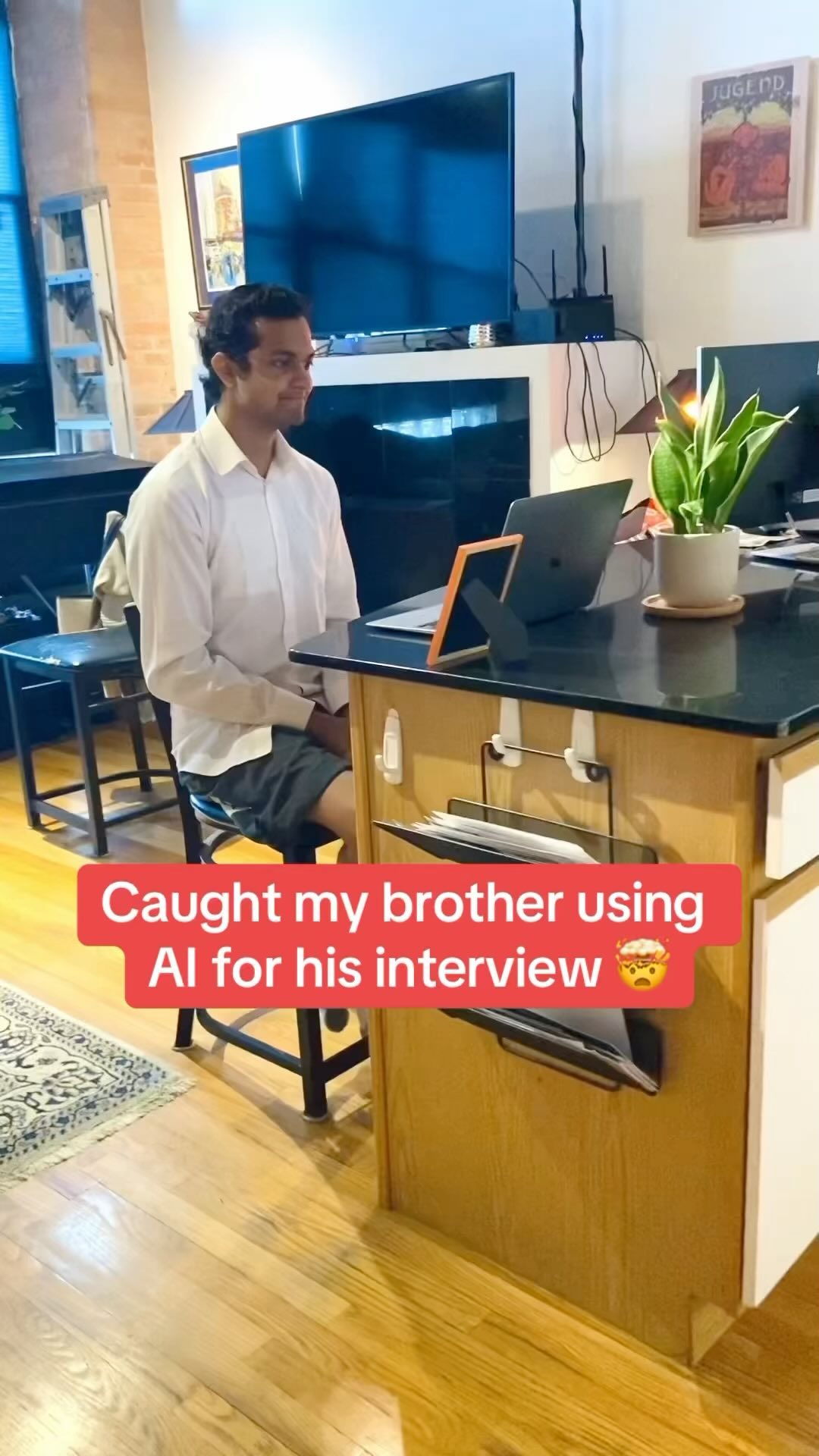 A man sits at a kitchen island, appearing to use a laptop in what looks like a home setting. A text overlay in a red box states, 'Caught my brother using AI for his interview' with an angry emoji.