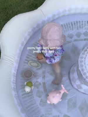 A high-angle shot captures a baby in a pink sun hat and swim diaper, submerged in a small, light-colored inflatable pool. The pool is filled with water and has various floating toys, including a pink fish. The background appears to be green grass.