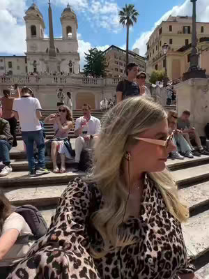 A woman with blonde hair and sunglasses poses for a selfie on a staircase. She is wearing a leopard-print blouse. In the background, there are buildings, stairs with people, and a clear sky. The overall composition is a selfie-style shot with a focus on the woman and the backdrop of a scenic urban location.