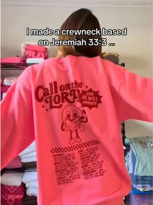 A person wearing a bright pink crewneck sweatshirt, with text and a graphic on the back that says "Call on the Lord and He will answer" near a cartoon style figure. The person is shown from the back, with brown hair, and is standing indoors. The background contains hints of shelves with items.