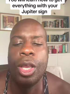 A man with a bald head and dark skin looks directly at the camera, with a slightly open mouth as if speaking. The background includes a bookshelf filled with books. Text at the top reads: 'You will learn how to get everything with your Jupiter sign.'