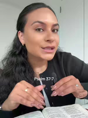 A woman with dark hair and fair skin is seen up close. She is wearing a black top and has a microphone clipped to her clothing. Her hands are clasped in front of her, and she is holding an open book, with text overlayed on the lower portion of the screen.