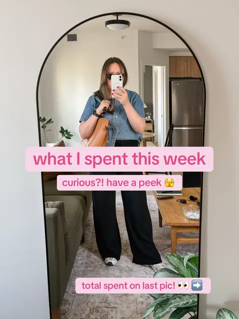A woman in a mirror selfie, standing in an apartment, holding a phone. She is wearing a denim shirt, black pants, and white sneakers. Text overlays on the image in pink rectangles. The background includes furniture and plants.
