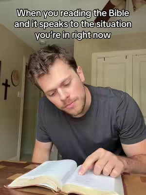 A medium shot of a man reading a Bible. The man is fair-skinned with short, brown hair and is wearing a dark grey t-shirt. He is seated at a wooden table, with the open Bible in front of him, and his right index finger pointing at the text. The background features a glimpse of a home interior, including a wall with a cross and an object resembling a wall clock or decorative element.