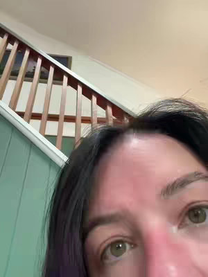 A close-up shot of a woman's face looking toward a staircase with a wooden bannister and green walls. Her skin is slightly flushed, and her eyes are open. The camera angle is from below.