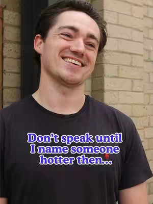 A man smiles broadly, wearing a black t-shirt with text overlaid in a blue and white stylized font. The background features a light-colored brick wall section.