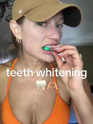 A selfie-style video cover image features a person holding a dental tool above their mouth. The person, centered in the frame, is wearing a neutral-colored baseball cap and an orange sports bra. Below the person, there is text that says "teeth whitening" with an emoji of a tooth and two hands forming a heart shape.