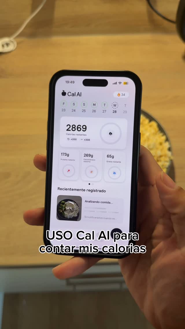 A person holding a smartphone displaying a diet tracking application. The screen shows the app interface with calorie and macronutrient information, alongside a partially visible meal image and a weekly calendar. The person, whose hand is visible, is holding the phone in front of a simple countertop. The background is slightly blurred with some yellow and a wood surface.
