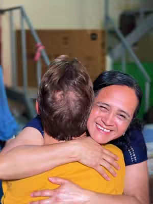 Two people are embracing, with the person in the foreground wearing a yellow shirt and the person in the background wearing a dark blue shirt. The background is slightly blurred, with glimpses of a metal structure and cardboard boxes.