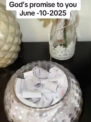 A close-up shot of a glass bowl filled with numerous rolled-up pieces of paper. The bowl sits on a black surface, which is also the background.  Behind the bowl, there's a decorative glass bottle with a lace sleeve and what looks like green stems and leaves inside. To the left of the bottle is another decorative white object.