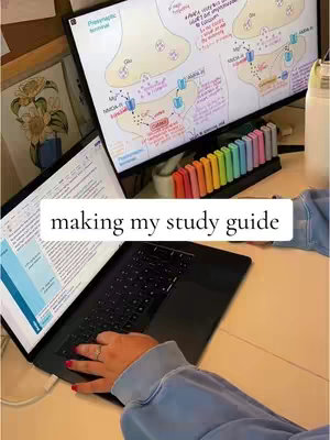The image shows a top-down view of a desk setup. A laptop is open, revealing a document on the screen. A second monitor displays a detailed diagram of a neuron. Colored highlighters are neatly arranged in a holder. A hand with red nail polish is actively using the laptop, indicating activity.