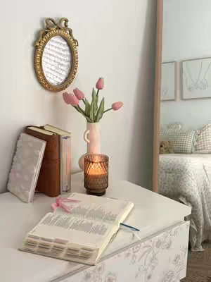 The image shows a well-lit bedroom scene with various objects arranged on a white surface, possibly a dresser or a table. On the left, a stack of books and a notebook are placed next to an ornate, golden-framed mirror hanging on the wall, reflecting a blurry view. In the center, a vase with pink tulips and a lit candle in a glass holder creates a warm ambience. An open book with a blue bookmark sits prominently, with a pink ribbon lying nearby. The background includes a mirror, part of a bed with pillows and a floral duvet implying a cozy and aesthetically pleasing environment.