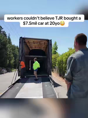 The image shows a scene of car transportation. The camera angle is medium, capturing a car carrier truck with a large loading area extending towards the viewer. Two men in work attire, one in an orange vest and the other in a neon green shirt, are standing near the vehicle. The backdrop features a road, trees, and a clear blue sky. The overall composition focuses on the workers in anticipation of the car’s arrival or loading.