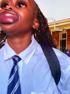 A close-up shot of a person's face with exaggerated features caused by a filter. The person has braided hair, wears a white collared shirt and tie, and is outside a building. The background is blurred.