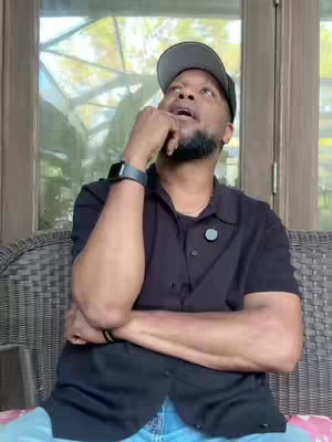 A man with a short beard and a black cap is seated on a woven chair, looking upwards with a contemplative expression. He is wearing a black button-down shirt and jeans. The background includes a window with foliage.