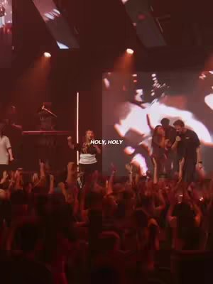 A live performance with a woman on stage, singing into a microphone, with a large screen in the background displaying abstract graphics. The audience is visible in the foreground with their arms raised, lit by red and orange stage lights.