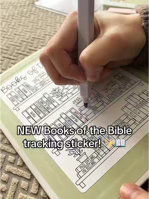 The image shows a hand holding a pen over a white sticker designed to graphically track the books of the Bible. The sticker is on top of a light green surface that resembles a notebook. The book titles are listed on shelves with floral decorations.