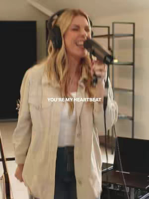A woman with long blonde hair is singing into a microphone in a recording studio. 
She is wearing headphones and a light-colored jacket over a white top. The background includes studio equipment and shelves.