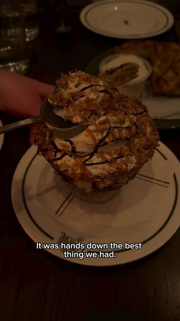 A close-up shot of a dessert, likely a sundae, being served. The sundae sits in a clear glass, layered with white cream, brown topping, and sprinkled with what appears to be toasted coconut flakes. A spoon is in the sundae, in the process of scooping a bite.