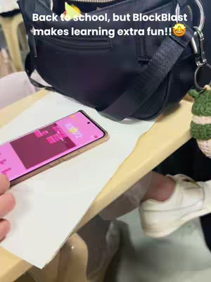 The image shows a phone on a table with a BlockBlast game on the screen. In the background there is a black purse and the legs of a person. 