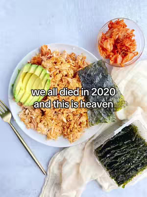 A top-down shot of a plate with shredded salmon-like food, a section of avocado slices, and a sheet of nori seaweed. To the right is a small glass bowl with a vibrant red, kimchi-like substance. A gold fork is on the table to the left. The background is a light, somewhat textured surface.