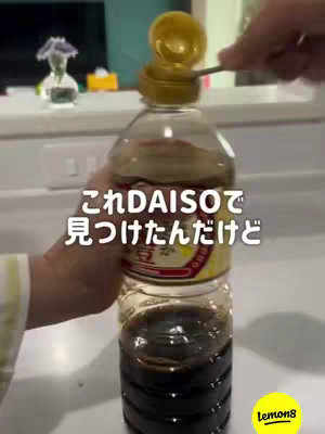 The image shows a soy sauce bottle with a yellow cap. A hand, with a yellow and white sleeve, is using a spoon to open the cap. White countertop and some decor can be seen in the background.