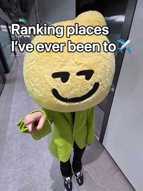 The image features a person wearing a large, yellow, mascot-style head resembling a smiling character. The person is also wearing a green blazer, black pants, and black shoes. The background is a hallway with a neutral-toned floor and walls. Text overlaid on the image reads, 'Ranking places I've ever been to,' with an airplane emoji.