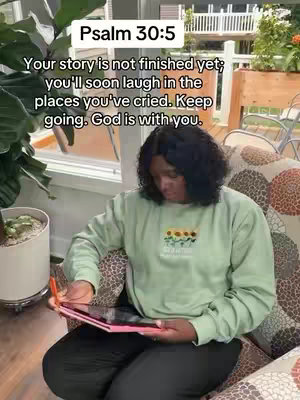 A person is sitting on a patterned armchair, wearing a green sweatshirt, and holding a pink tablet. The background shows an indoor setting with a window and plants, with additional text overlay.