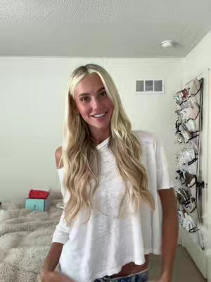 A young woman with long blonde hair smiles at the camera, wearing a white off-the-shoulder top and dark jeans. She stands in a room with a bed in the background to the left and a wall with a vent/air conditioner above. The room is lit with natural light.